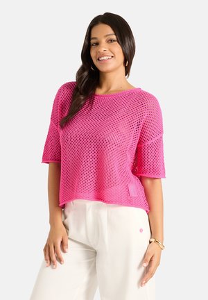 Woman with long black hair smiles wearing a bright pink open-knit short sleeve top and white pants against a plain light background.