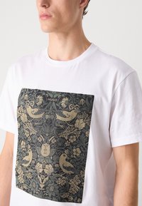 Weißes Baumwoll-T-Shirt mit einem großen schwarzen und goldenen Blumenmuster mit Vögeln, zentral auf der Vorderseite, mit der Aufschrift "HUGO" oben.