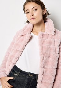 Giacca in pelliccia sintetica di colore rosa chiaro con un design trapuntato. Collo alto, texture morbida e chiusura con bottoni. Indossata sopra una camicia bianca e jeans scuri.