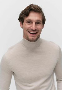 Beige coltrui gemaakt van zachte stof, met een aansluitend ontwerp en geribbelde hals, gedragen door een glimlachende persoon tegen een effen achtergrond.