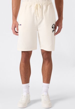 Mann trägt off-white Shorts mit Kordelzug, kleinem MLB-Logo am linken Oberschenkel und schwarzem Schriftzug am rechten Oberschenkel, weiße Socken und weiße Sneakers.