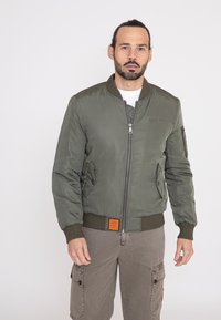 Olivengroene bomberjack met een ritssluiting aan de voorkant, ribgebreide manchetten en een ribgebreide taille. Bevat twee zijzakken en een oranje label bij de zoom. Glad van textuur.