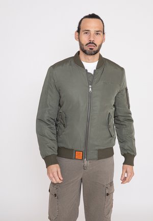 Olive green bomber jacket with a zip front, ribbed cuffs, and waist. Features two side pockets and an orange label at the hem. Smooth texture.