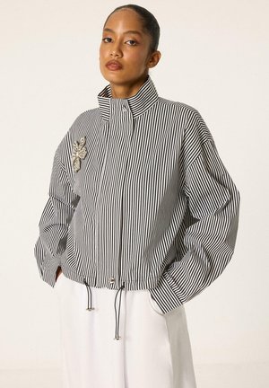 Femme aux cheveux tirés en arrière porte une veste à col haut rayée verticalement noir et blanc avec une broche florale et un pantalon blanc, mains dans les poches.