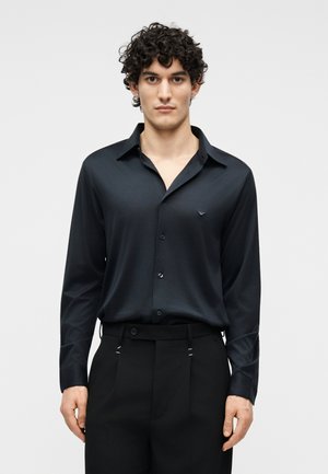 Νέος άνδρας με σγουρά μαλλιά, που φοράει μαύρο πουκάμισο με μακριά μανίκια και κουμπιά, το οποίο είναι μπα tucked μέσα σε μαύρο παντελόνι με ψηλή μέση, στέκεται μπροστά σε απλό φόντο.