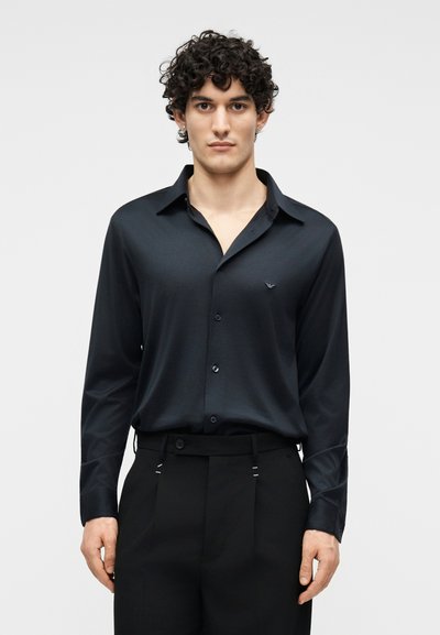 Jeune homme aux cheveux bouclés portant une chemise noire à manches longues boutonnée, rentrée dans un pantalon noir taille haute, debout devant un fond uni.