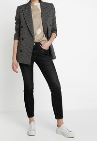 Person som bär en svart och vit rutiga kavaj, en beige mönstrad skjorta, svarta tajta jeans och vita sneakers, stående med en hand i fickan.