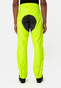 Pantaloni impermeabili gialli brillanti con una toppa circolare nera, vita elastica, zip laterali e dettagli riflettenti sui bordi.