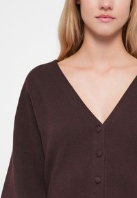 Cardigan bordeaux con scollatura a V profonda, tre bottoni sulla parte anteriore e tessuto morbido e strutturato. Presenta maniche larghe e sgonfie.