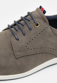 Chaussure en suede gris avec lacets gris, œillet doré et accents cousus. Le motif latéral comprend de petites perforations. Semelle extérieure blanche.