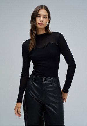 Mujer con cabello largo y castaño lleva una blusa transparente negra de manga larga con detalle de ondas con cuentas y pantalones de cuero negros de tiro alto contra un fondo gris.