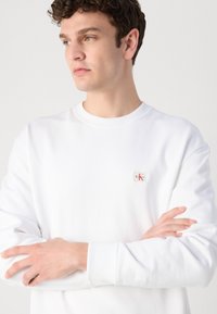 Sudadera blanca en mezcla de algodón, con cuello redondo, puños acanalados y un pequeño parche con logo en el pecho. Textura suave, diseño casual.