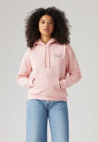 Rosa bomullshoodie med frampocket, dragsko i huvan och en liten texturerad logotyp. Matchad med ljusblå vida jeans.