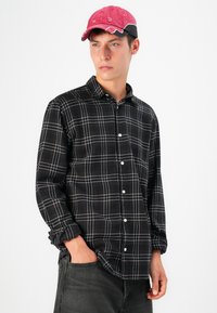 Camisa de franela a cuadros en blanco y negro con mangas largas y cierre de botones, combinada con una gorra rosa y negra, llevada con jeans grises.