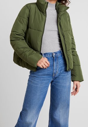 Persoon draagt een opengewerkte olijfgroene donsjas over een grijze geribde trui en blauwe hooggelaagde wijde jeans tegen een effen achtergrond.