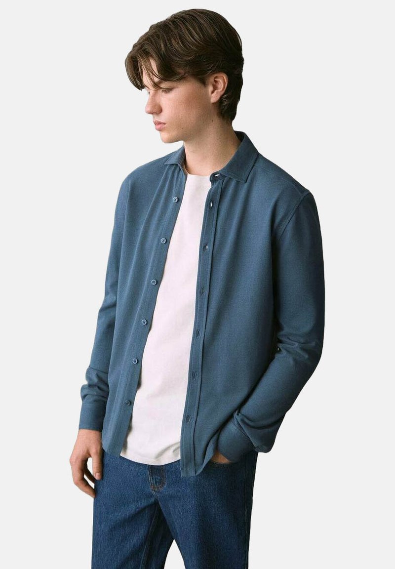 Giovane uomo con capelli castani che indossa una camicia button-up turchese sopra una maglietta bianca e jeans blu, guarda in basso con una mano in tasca.