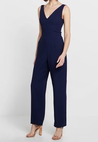 Mörkblå byxdress med djup V-ringning, ärmlös design, skräddarsydd passform och vida ben. Har diskreta veck och en mjuk textur.