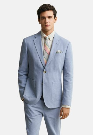 Homme portant un costume bleu clair avec une veste boutonnée, une chemise blanche, une cravate à rayures pastel et un mouchoir de poche, debout avec une main dans la poche.