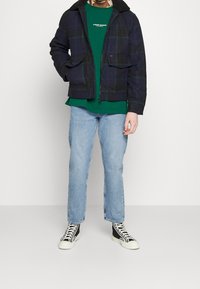 Marinblå rutig jacka med svart pälsfodrad krage, kombinerad med en grön t-shirt och ljusblå raka jeans. Svarta högskaftade sneakers.