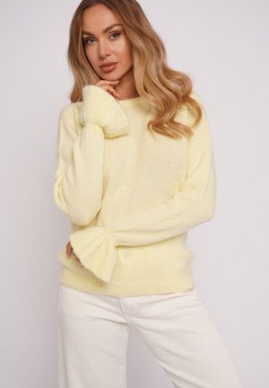 Kobieta o długich falowanych włosach, ubrana w żółty puszysty sweter z rozkloszowanymi mankietami i białe spodnie, pozująca z jedną ręką przy twarzy.