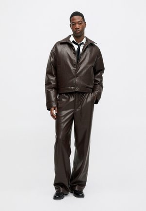 Ruda odinė trumpa striukė su dideliu apykakle ir mygtukų uždarymu, derinama su atitinkamomis plačiomis kelnėmis ir juodais batais.