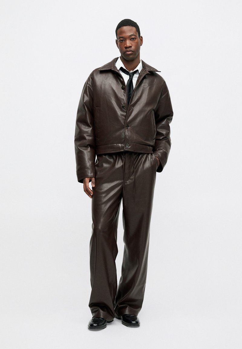 Jachetă scurtă din piele maro, cu guler mare și închidere cu buton, asortată cu pantaloni largi de aceeași culoare și pantofi negri.