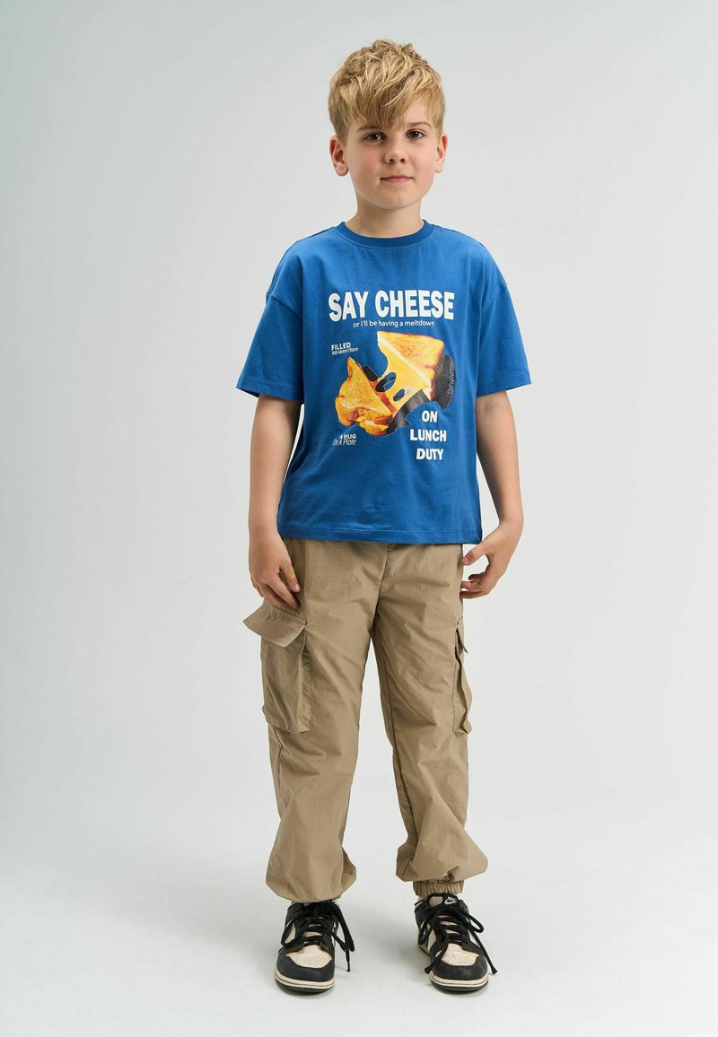 Blå T-shirt med teksten "Say Cheese" og grafik af ost på; beige cargobukser med sidelommer; sorte sneakers med hvide detaljer.