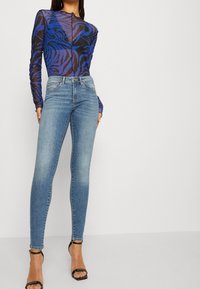 Long-sleeve bodysuit in sheer black with blue tiger stripes, paired with light-wash skinny jeans. Black stiletto heels complete the outfit.
