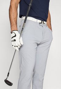 Γκολφ ενδυμασία που περιλαμβάνει γκρι υφασμάτινα παντελόνια, μια λευκή ζώνη με μεταλλική πόρπη και ένα μαύρο και λευκό γάντι γκολφ, κρατώντας μια ρακέτα.