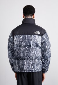 Pufferjakke i sort og hvidt abstrakt mønster med en ensfarvet sort øvre sektion og høj krave. Har logo på ryggen.