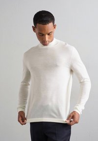Maglietta bianca a maniche lunghe con collo alto, in tessuto a maglia, design aderente e polsini a coste. Indossata sopra pantaloni blu navy. Aspetto leggermente testurizzato.