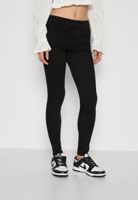 Sorte skinny jeans med en glatt tekstur og klassisk femlommersdesign, båret med svarte og hvite sneakers og en hvit bluse.