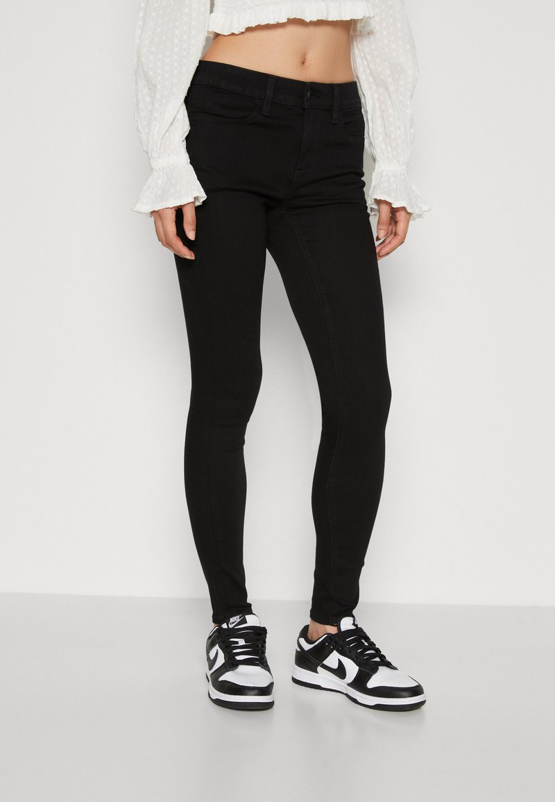Sorte skinny jeans med en glatt tekstur og klassisk femlommersdesign, båret med svarte og hvite sneakers og en hvit bluse.