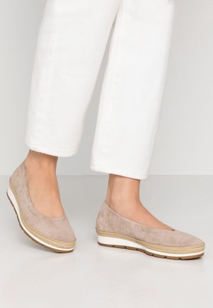 Beżowe zamszowe buty wsuwane z zaokrąglonym noskiem, ozdobione plecioną lamówką i kontrastującą białą podeszwą na platformie. Widoczne białe spodnie do kostek.