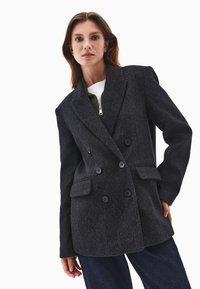 Tummanharmaa kaksirivinen bleiseri, joka on valmistettu teksturoidusta kankaasta. Siinä on suuret napit, viistot läpät ja kaksi etutaskua.