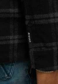 Gros plan d’une manche de chemise à carreaux noirs et gris avec un bouton noir et une étiquette en tissu "CHASIN'" au-dessus d’un jean bleu.