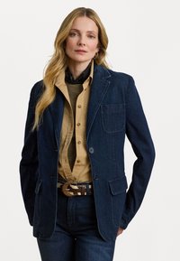 Donkerblauwe denim blazer met notch-kraag, twee voorzakken, knoopsluiting en een gestructureerd inner shirt. Een bruine met patroon riem maakt de look compleet.
