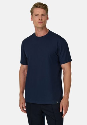 Mężczyzna o krótkich blond włosach, ubrany w gładki granatowy t-shirt z krótkim rękawem i ciemne spodnie, stojący na jasnym tle.