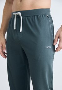 Muške tamnozelene trenirke od mekog materijala, s pojasom na vezanje, bočnim džepovima i bijelim "BOSS" logotipom na lijevoj nozi.