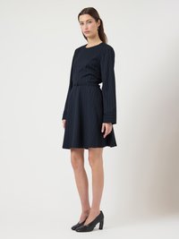 Γυναίκα που στέκεται φορώντας φορέματα με ναυτικό μπλε ρίγα, μακρυμάνικο και ζώνη, με μαύρες ψηλοτάκουνες γόβες, μπροστά σε ένα απλό λευκό φόντο.