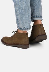 Botas de cuero marrón hasta el tobillo con punta redondeada, diseño de cordones y suela de goma. Presentan sutiles acentos de costura y una textura suave.