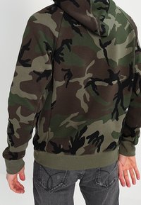 Maskáčová mikina v odstínech zelené a hnědé, vyrobená z měkké tkaniny, s kapucí, žebrovanými manžetami a přední kapsou ve stylu klokan.