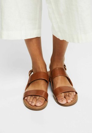 Persona con pantalones blancos cortos y sandalias de cuero marrón con dos tiras anchas sobre pies desnudos sobre fondo blanco.