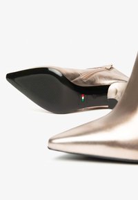 Metallisch roségoldene High-Heels mit spitzem Zeh, glatter Textur und einer schwarzen Gummisohle mit strukturiertem Grip und Reißverschlusdetail.