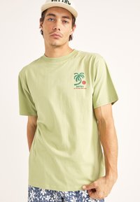 Camiseta de algodón verde claro con mangas cortas, cuello redondo y un gráfico de palmera con un círculo rojo en el lado izquierdo del pecho.