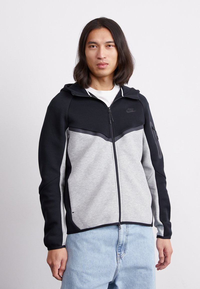 Nike zip-up kapuca v črni in sivi barvi. Ima kapuco, kontrastne panele in žep na zadrgo. Mehka tekstura, dizajn športne oblačila in prilegajoča silhueta.