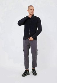 Black zip-up jacket with textured fabric and fitted design, paired with grey cargo pants featuring side pockets and black sneakers.
