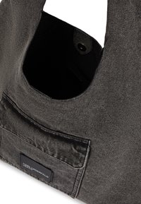 Bolsa de denim cinza com bolso frontal e etiqueta preta com a inscrição "Karl Lagerfeld Jeans," parte superior aberta com fecho de botão de pressão revelando interior preto.