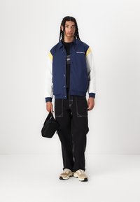 Veste bleu marine avec des accents blancs et jaunes, associée à un pantalon noir et des baskets beiges. Sac noir tenu à la main gauche.