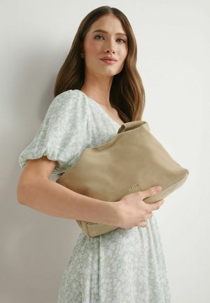 Młoda kobieta w jasnooliwkowej sukience w kwiaty trzyma dużą beżową zamszową kopertówkę z wytłoczonym logo na jednolitym tle.